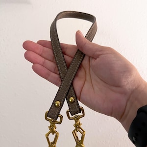 May include: A pair of taupe-colored leather bag straps with gold-toned metal hardware. The straps are stitched with a lighter thread and feature swivel clasps for easy attachment. The hardware includes rivets and swivel clasps.