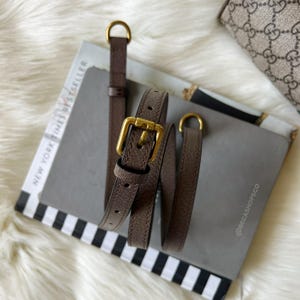 May include: A brown leather belt with a gold-colored buckle and D-ring hardware. The belt is coiled on top of a book with the words "NEW YORK TIMES BESTSELLER" visible. The background includes a patterned book and a fluffy white surface.