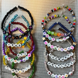 May include: A set of 10 colorful beaded bracelets with white letter beads spelling out the names of Taylor Swift albums: "Debut", "Fearless", "Speak Now", "Red", "1989", "Reputation", "Lover", "Folklore", "Evermore", and "Midnights".