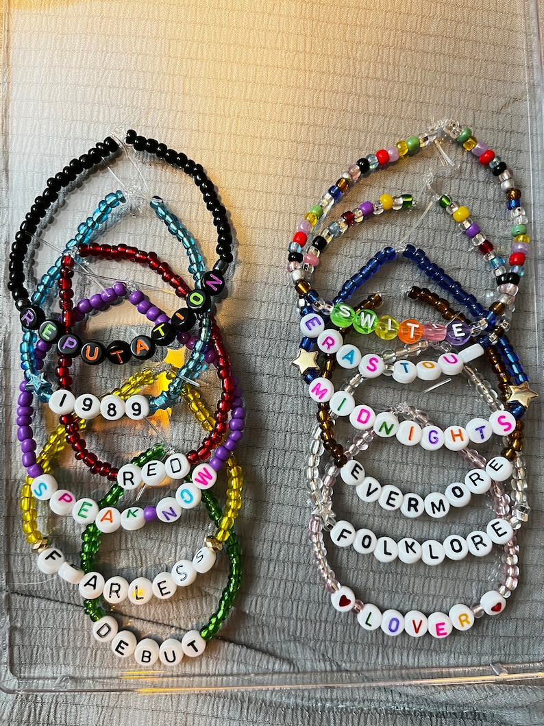 May include: A set of 10 colorful beaded bracelets with white letter beads spelling out the names of Taylor Swift albums: "Debut", "Fearless", "Speak Now", "Red", "1989", "Reputation", "Lover", "Folklore", "Evermore", and "Midnights".