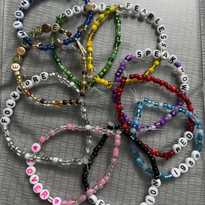 May include: A collection of colorful beaded bracelets with different words spelled out on them, including "Fearless", "Speak Now", "Red", "1989", "Reputation", "Lover", "Folklore", "Evermore", "Delicate", and "Reputation".