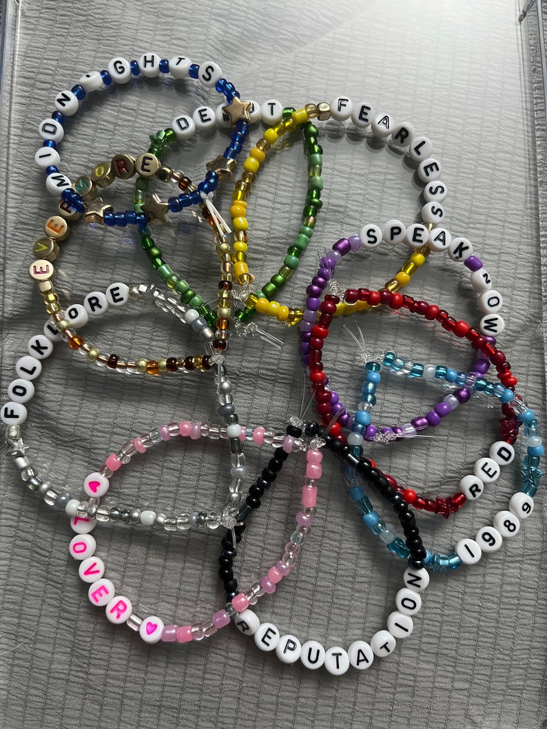 May include: A collection of colorful beaded bracelets with different words spelled out on them, including "Fearless", "Speak Now", "Red", "1989", "Reputation", "Lover", "Folklore", "Evermore", "Delicate", and "Reputation".