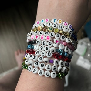 May include: A stack of white beaded bracelets with different colored letters spelling out the names of Taylor Swift albums: "Reputation", "Lover", "1989", "Red", "Speak Now", "Fearless", "Debut", "Folklore", "Lover", "Reputation", and "Taylor Swift".