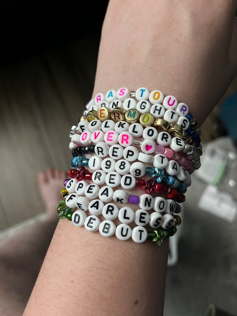 May include: A stack of white beaded bracelets with different colored letters spelling out the names of Taylor Swift albums: "Reputation", "Lover", "1989", "Red", "Speak Now", "Fearless", "Debut", "Folklore", "Lover", "Reputation", and "Taylor Swift".