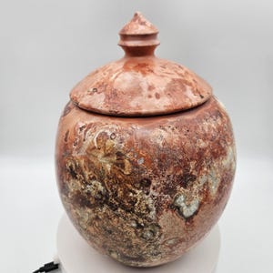 May include: A round, lidded vessel featuring a mottled, textured surface with variegated shades of reddish-brown, tan, and dark brown. The lid is domed and topped with a small, pointed finial, matching the vessel's earthy, marbled appearance.
