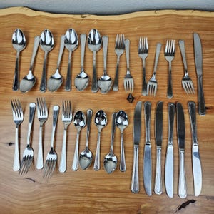 May include: A collection of stainless steel flatware including various spoons, forks, and knives arranged in two rows. The set features polished metal surfaces with simple, smooth handles, showcasing a variety of sizes and functional shapes.