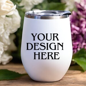 Peut inclure: Un gobelet &agrave; vin en acier inoxydable blanc avec un couvercle noir et le texte "YOUR DESIGN HERE" imprim&eacute; sur le c&ocirc;t&eacute;. Le gobelet est pos&eacute; sur une surface en bois avec des fleurs blanches et violettes en arri&egrave;re-plan.
