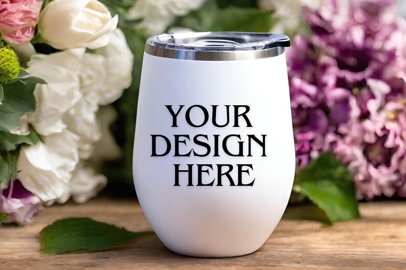 Peut inclure: Un gobelet &agrave; vin en acier inoxydable blanc avec un couvercle noir et le texte "YOUR DESIGN HERE" imprim&eacute; sur le c&ocirc;t&eacute;. Le gobelet est pos&eacute; sur une surface en bois avec des fleurs blanches et violettes en arri&egrave;re-plan.