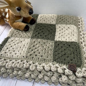 May include: A handmade crochet blanket in a patchwork design, featuring squares of beige, olive green, and light green. The blanket has a scalloped edge and a small brown tag. A stuffed deer toy rests on the blanket.