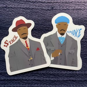 May include: Two stickers featuring illustrations of men in suits. One wears a red hat and has the word "$TAGK" next to him. The other wears a blue cap and has the word "SMOKE" next to him.