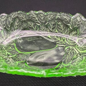 May include: A green glass dish with a leaf design. The dish has two handles and a shallow, oval shape. The leaf pattern is embossed on the sides and bottom of the dish. The glass has a slight glow.