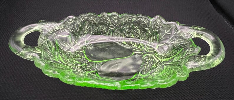 May include: A green glass dish with a leaf design. The dish has two handles and a shallow, oval shape. The leaf pattern is embossed on the sides and bottom of the dish. The glass has a slight glow.