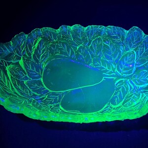 May include: A glowing green glass serving dish with a leaf and pear design. The oval dish has two handles and is illuminated with a black light, showcasing its intricate details and vibrant color.