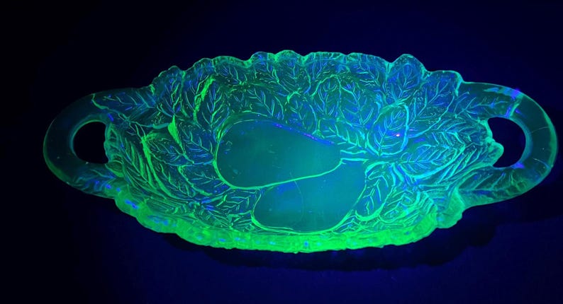 May include: A glowing green glass serving dish with a leaf and pear design. The oval dish has two handles and is illuminated with a black light, showcasing its intricate details and vibrant color.