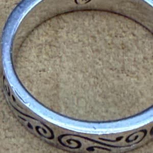 May include: A silver-toned metal ring featuring a continuous black engraved swirl pattern around the outer band. The interior of the ring band displays a small, stamped heart-shaped hallmark symbol centered at the top of the inner circumference.
