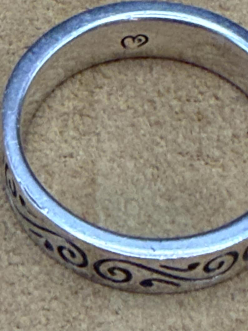May include: A silver-toned metal ring featuring a continuous black engraved swirl pattern around the outer band. The interior of the ring band displays a small, stamped heart-shaped hallmark symbol centered at the top of the inner circumference.
