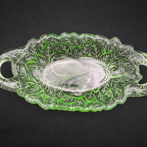 May include: A vintage, oval-shaped, green glass serving dish with a leaf design. The dish has two handles and a scalloped edge. The leaf pattern is visible throughout the dish, adding a decorative touch. The dish is likely used for serving food.