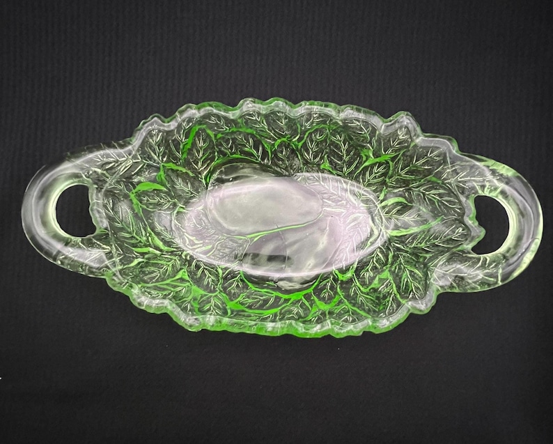 May include: A vintage, oval-shaped, green glass serving dish with a leaf design. The dish has two handles and a scalloped edge. The leaf pattern is visible throughout the dish, adding a decorative touch. The dish is likely used for serving food.