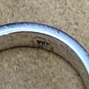 May include: A silver-colored metal ring with a smooth, polished surface. The interior of the band features a small, stamped hallmark that reads 925, indicating the metal composition. The ring has a simple, rounded profile and a reflective finish.
