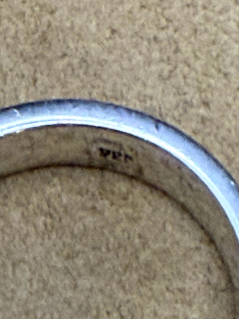 May include: A silver-colored metal ring with a smooth, polished surface. The interior of the band features a small, stamped hallmark that reads 925, indicating the metal composition. The ring has a simple, rounded profile and a reflective finish.
