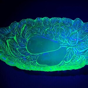 May include: A glowing green glass serving dish with a leaf and pear design. The dish has two handles and is illuminated with a black light, creating a vibrant, eye-catching display. The dish is likely vintage.