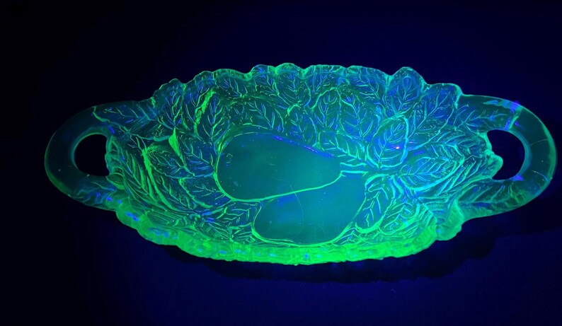May include: A glowing green glass serving dish with a leaf and pear design. The dish has two handles and is illuminated with a black light, creating a vibrant, eye-catching display. The dish is likely vintage.
