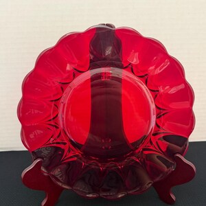 May include: A vibrant ruby red glass plate featuring a smooth circular center and a wide, scalloped rim with deep, curved indentations. The translucent glass catches the light, creating varying shades of red across the textured surface.