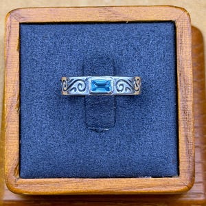 May include: A silver ring featuring a rectangular, light blue gemstone set in the center. The band is decorated with engraved, swirling scrollwork patterns that extend across the surface on both sides of the central stone setting.