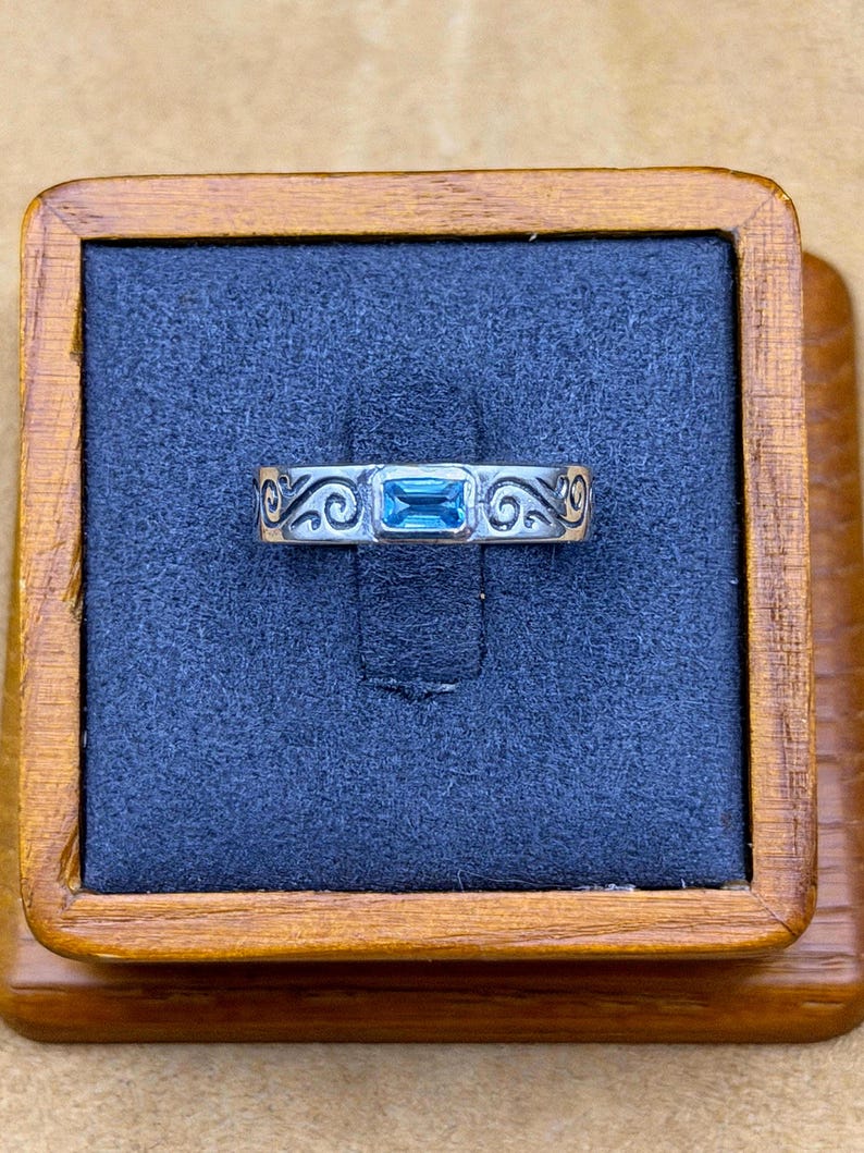May include: A silver ring featuring a rectangular, light blue gemstone set in the center. The band is decorated with engraved, swirling scrollwork patterns that extend across the surface on both sides of the central stone setting.