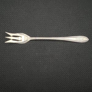 May include: A silver fork with three tines and a long handle. The handle has a decorative floral design near the end. The fork is likely for serving appetizers or small portions.