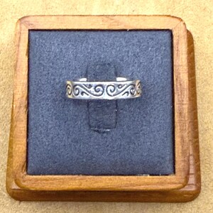 May include: A silver-toned metal ring featuring an embossed pattern of repeating swirls along the outer band. The ring is displayed on a dark gray square insert held within a square wooden frame with a natural light brown finish.