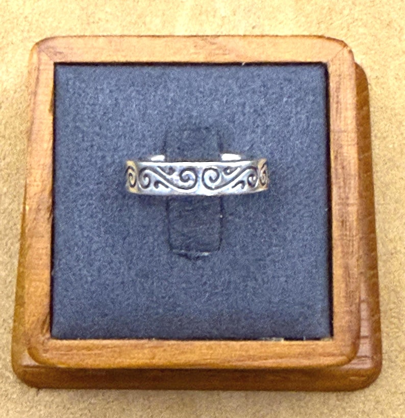 May include: A silver-toned metal ring featuring an embossed pattern of repeating swirls along the outer band. The ring is displayed on a dark gray square insert held within a square wooden frame with a natural light brown finish.