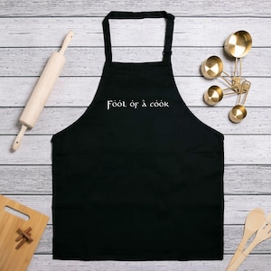May include: Black apron with the text "Fool of a Cook" in white script. The apron is displayed on a light wood-grain surface, alongside a rolling pin, measuring spoons, a cutting board, and wooden spoons.