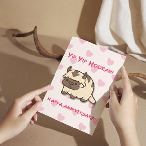 Puede incluir: Tarjeta de felicitación con una ilustración de dibujos animados de un personaje bisonte, rodeada de corazones rosas. La tarjeta dice "Yip Yip Hooray!" y "Happy Anniversary!"