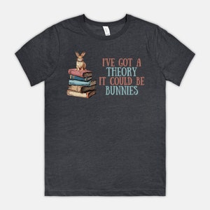 May include: Dark grey t-shirt featuring a bunny on a stack of books. Text reads "I've got a theory it could be bunnies" in coral and teal. A casual tee for those who love books and bunnies.