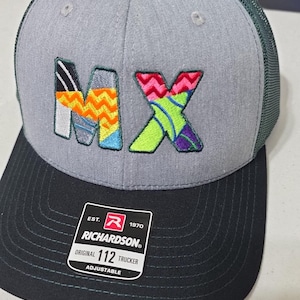 Può includere: Cappellino da camionista grigio e nero con le lettere "MX" ricamate in disegni colorati. Il cappello presenta una visiera nera, un pannello frontale grigio e un retro in rete verde. L'etichetta del marchio Richardson è visibile sul davanti.