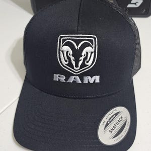 May include: Black and white embroidered Ram logo on a black trucker hat with a black mesh back. The hat has a white "The Authentic Yupoong Snapback Since 1974" sticker on the back.
