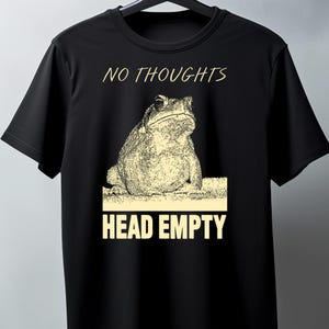Può includere: T-shirt nero con un'illustrazione beige di un rospo. Il testo "NO THOUGHTS" è sopra il rospo e "HEAD EMPTY" sotto. Il disegno è centrato sulla parte anteriore della maglietta.