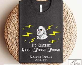 May include: Dark gray t-shirt featuring a white illustration of Benjamin Franklin wearing sunglasses, surrounded by yellow lightning bolts. The text reads "It's Electric Boogie Woogie Woogie" and "Benjamin Franklin June 10. 1752."