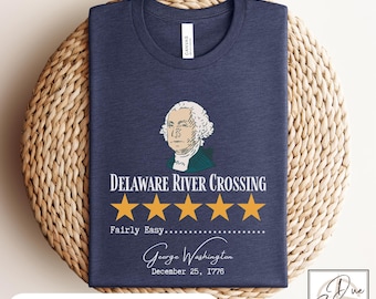 May include: Navy blue t-shirt featuring a graphic of George Washington and the text "Delaware River Crossing." The design includes five gold stars and the phrase "Fairly Easy." The shirt also displays the date December 25, 1776.