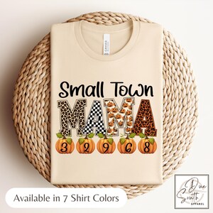 May include: A beige t-shirt with the words "Small Town MAMA" in a mix of leopard print, checkered, and pumpkin patterns. Below the text are orange pumpkins with numbers. The shirt is displayed on a woven surface. The text "Available in 7 Shirt Colors" is at the bottom.