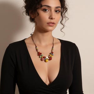 Puede incluir: Un collar de cuentas de colores con un cordón marrón. El collar presenta un grupo de cuentas de varios tamaños en tonos de rojo, amarillo, verde, blanco y rosa. La mujer de la imagen lleva una camiseta negra de manga larga.
