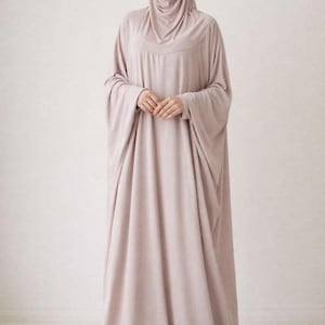 May include: A light mauve-colored modest dress with a matching hijab. The dress features long, flowing sleeves and a loose, comfortable fit. The fabric drapes elegantly, creating a modest and stylish silhouette. The outfit is suitable for various occasions.