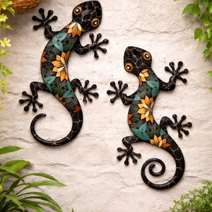 May include: Two metal gecko wall decorations. Each gecko is black with a mosaic design in teal, orange, and yellow. They are mounted on a textured, off-white wall. Green plants surround the geckos.