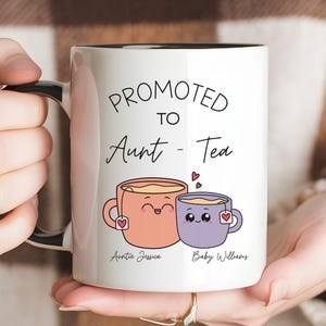 May include: A white ceramic mug with a black handle and interior. The front features the text "PROMOTED TO Aunt - Tea" above a graphic of two smiling tea cups with tea bags. Below the cups, the names "Auntie Jessica" and "Baby Williams" appear.