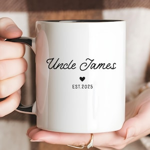 May include: White ceramic mug with a black interior and handle. The mug features the text "Uncle James" in a stylish script, a small heart, and the year "EST.2025" in a simple font. The mug is held by a person.