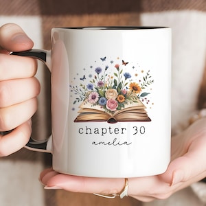 May include: White ceramic mug with a black interior, featuring a floral design above an open book. The text "chapter 30 amelia" is below the book. The mug is held by a person.