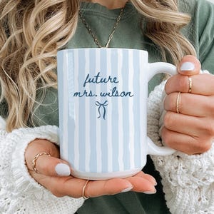 May include: A white ceramic mug featuring vertical light blue brushstroke stripes. The text "future mrs. wilson" is printed in a dark blue script font, positioned above a small, simple dark blue line drawing of a bow centered on the mug.