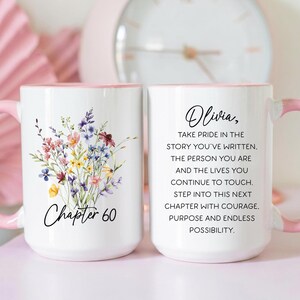 May include: White ceramic mug with a pink interior and handle. One mug features a floral design and the text "Chapter 60." The other mug has the name "Olivia" and inspirational text. A pink clock is in the background.