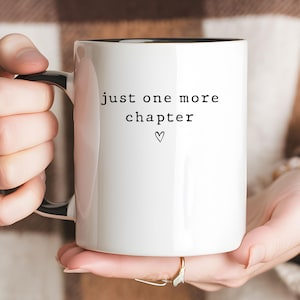 May include: A white ceramic mug with a black interior and handle, featuring the black text "just one more chapter" printed on the front, accompanied by a small, simple black heart symbol centered below the text in a clean, minimalist font.